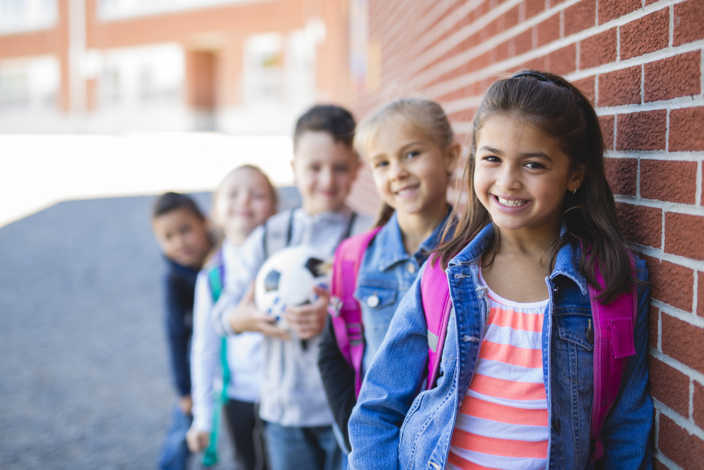 Élèves du primaire qui sont accotés sur une école, souriants. Un a un ballon dans ses mains.