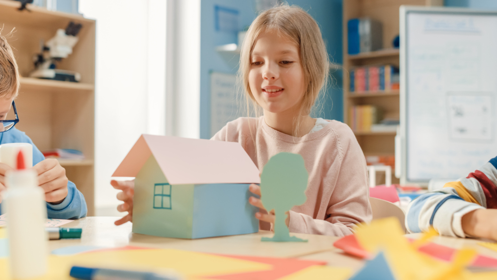 Elementary School Arts & Crafts Class: Cute Little Blond Girl Has Fun with Her Classmates on the Handicraft Project, Uses Colorful Paper, Glue a House